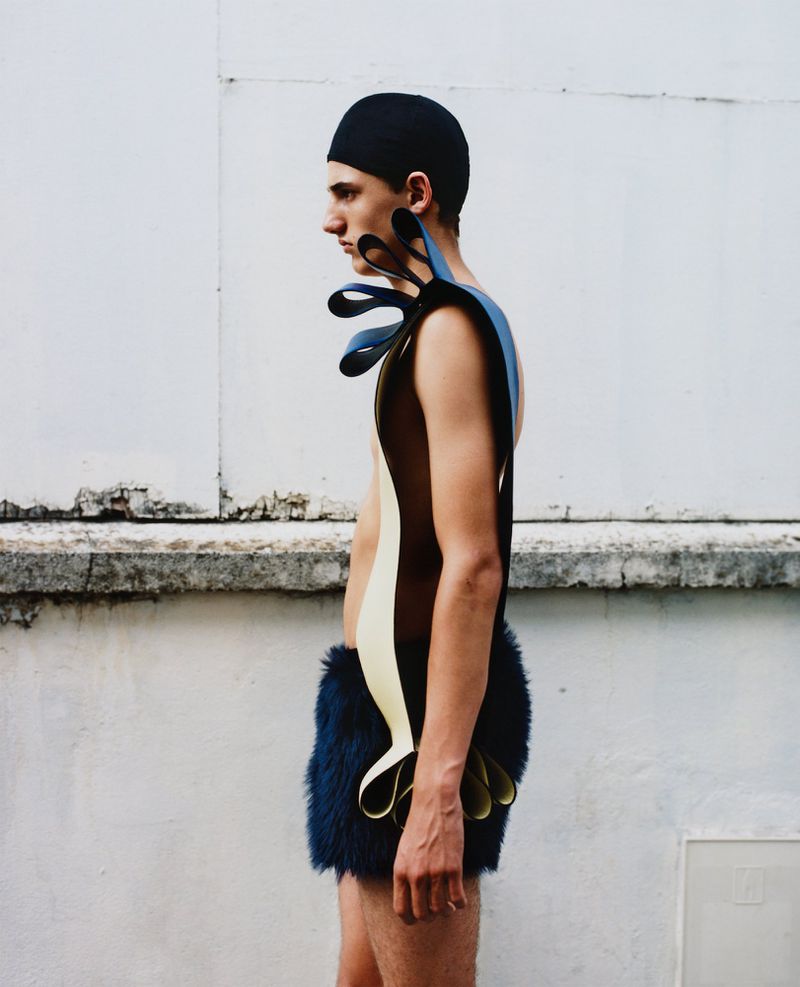 A slender male model in a cap and avant-garde outfit stands sideways against a white wall with a concrete ledge in an outdoor fashion shot.