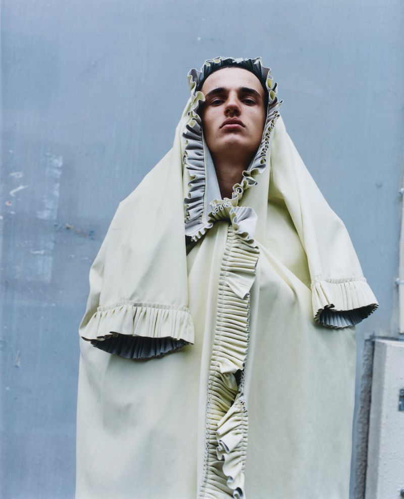 Adult male in pale yellow hooded robe with ruffled trim stands against a grey wall in soft lighting, looking directly at the camera.