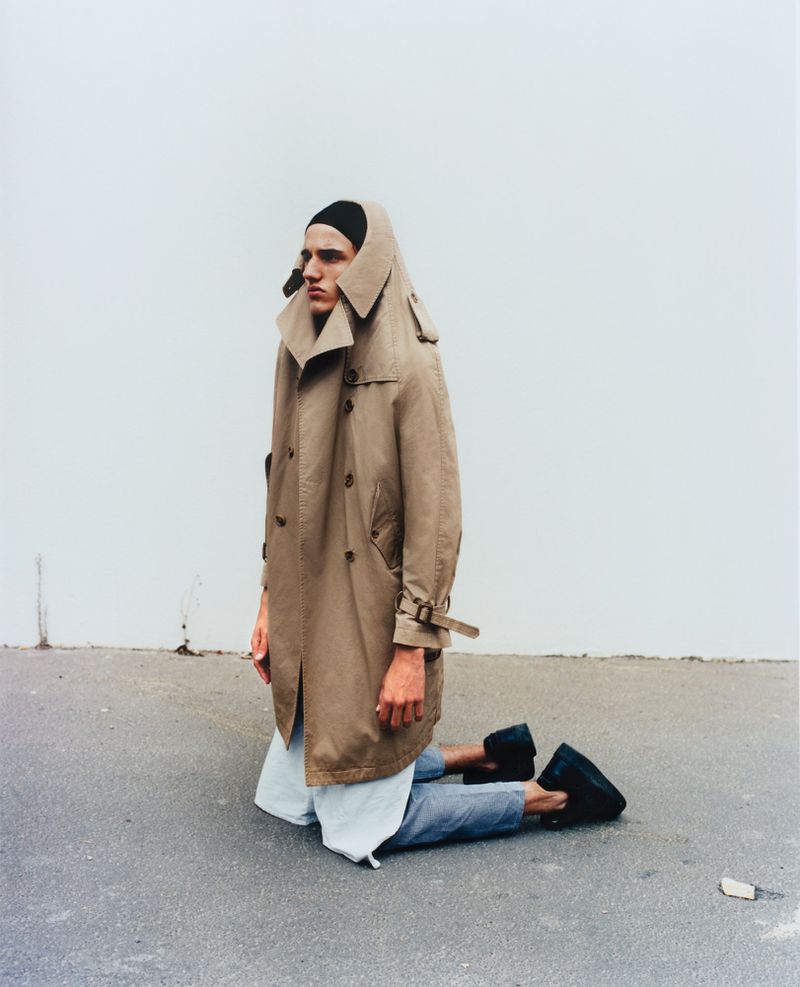 An adult male kneeling outdoors wearing a tan trench coat, black beanie, and light blue jeans against a white wall background.