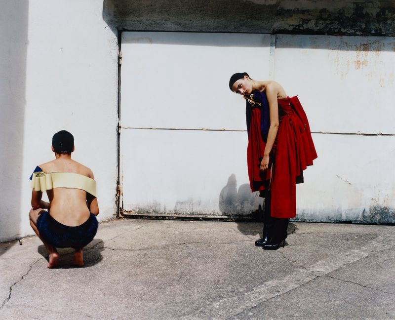 Fashion shot of a model standing with a red dress and a person crouching with a shoulder wrap, outside against a concrete wall.