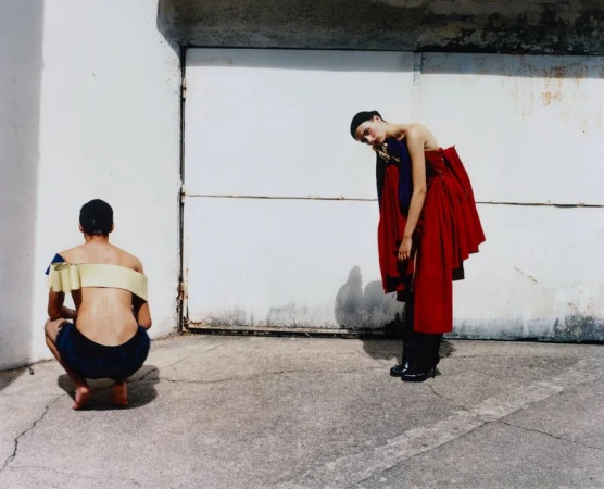 Fashion shot of a model standing with a red dress and a person crouching with a shoulder wrap, outside against a concrete wall.