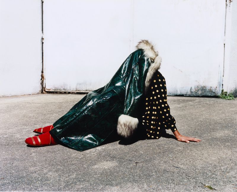 Person in an emerald-green vinyl coat with fur trim and red shoes posed on concrete in front of a white concrete wall.
