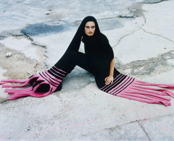 Model seated on concrete wearing a black hooded garment with exaggerated long sleeves ending in fringe and pink stripes.