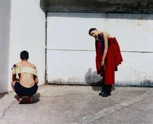 Two models outdoors against a white wall; one crouching in dark shorts and cream bandeau, another standing in a voluminous red dress.