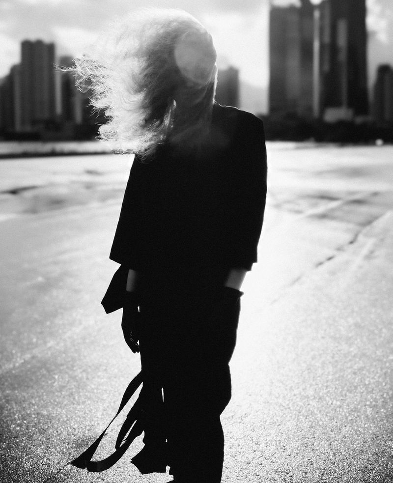 Monochrome portrait of a solitary adult woman stands on a beach, hair blowing, wearing a coat with a cityscape in the background.