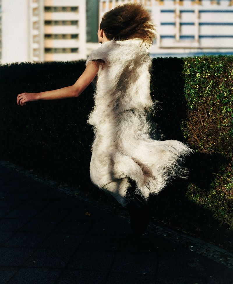 Woman in a unique white furry fashion dress, captured outdoors against a hedge and city backdrop, showcasing an editorial style.