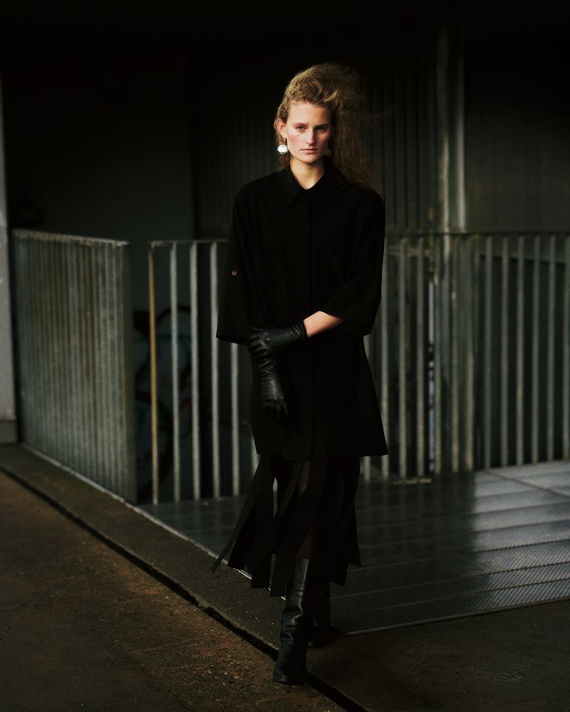 Fashionable woman in a black coat, skirt, and leather gloves stands in an urban location with wet pavement and a dark industrial background.