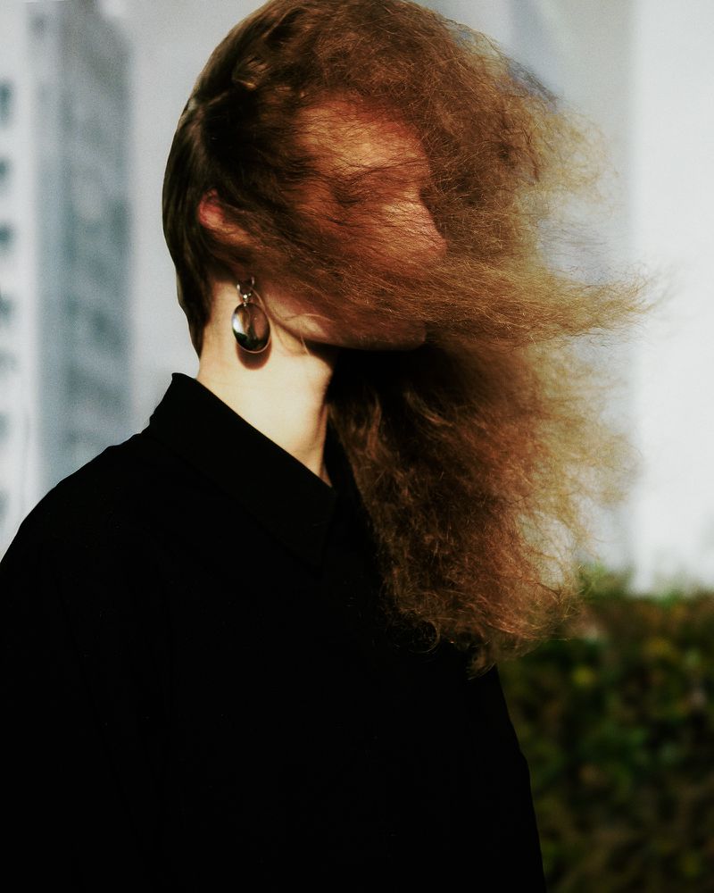Headshot of an adult female with long wavy brown hair obscuring her face, wearing a black top and large earrings, with city buildings blurred in background.