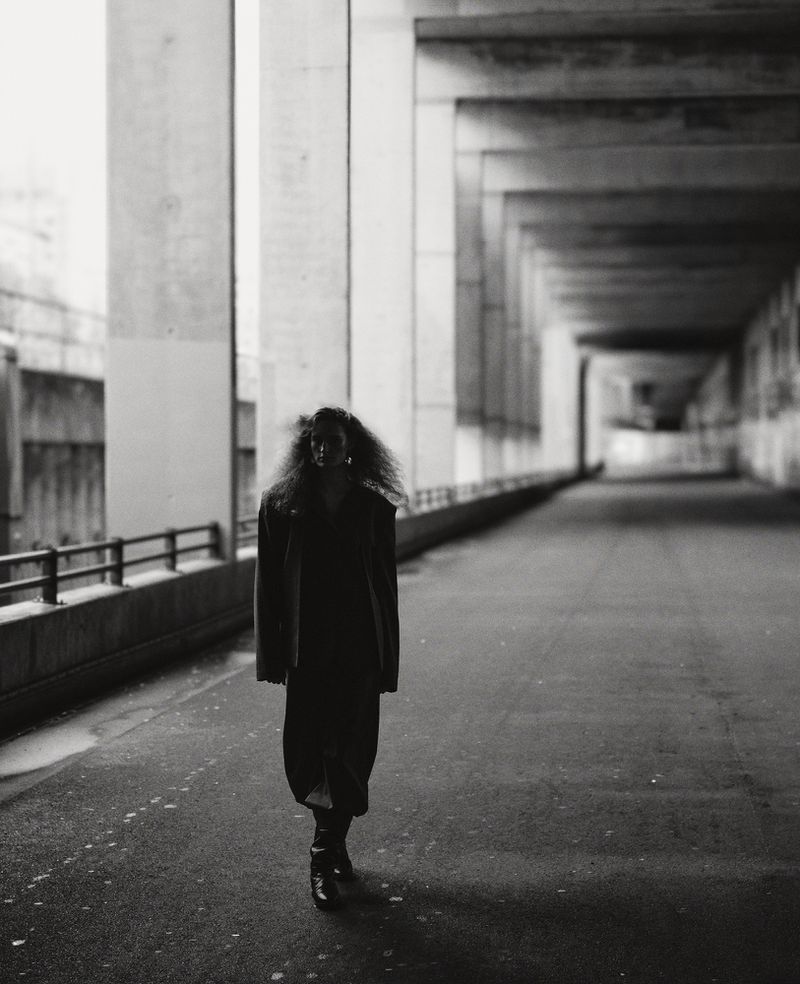 Monochrome shot of a woman with large curly hair walking under a concrete structure. She wears a dress and boots in an urban setting.
