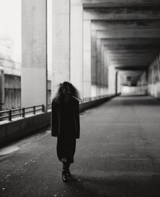 Monochrome shot of a woman with large curly hair walking under a concrete structure. She wears a dress and boots in an urban setting.