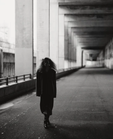 Monochrome shot of a woman with large curly hair walking under a concrete structure. She wears a dress and boots in an urban setting.