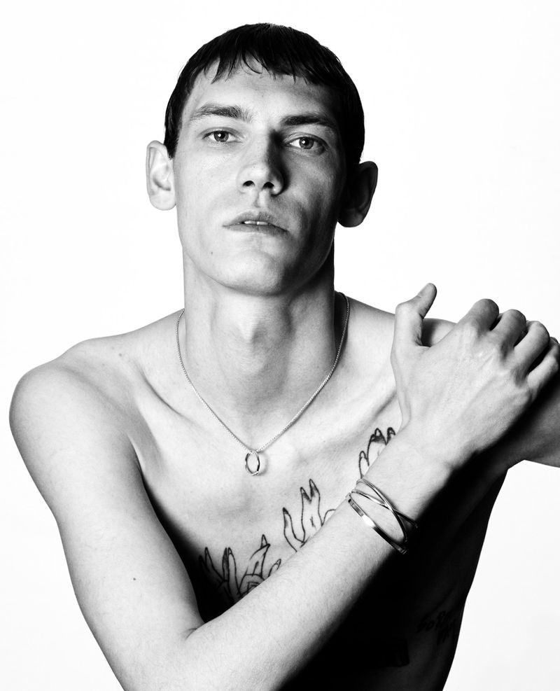 Black and white studio shot of a shirtless male model with necklace and bracelet, neutral facial expression, and light background.
