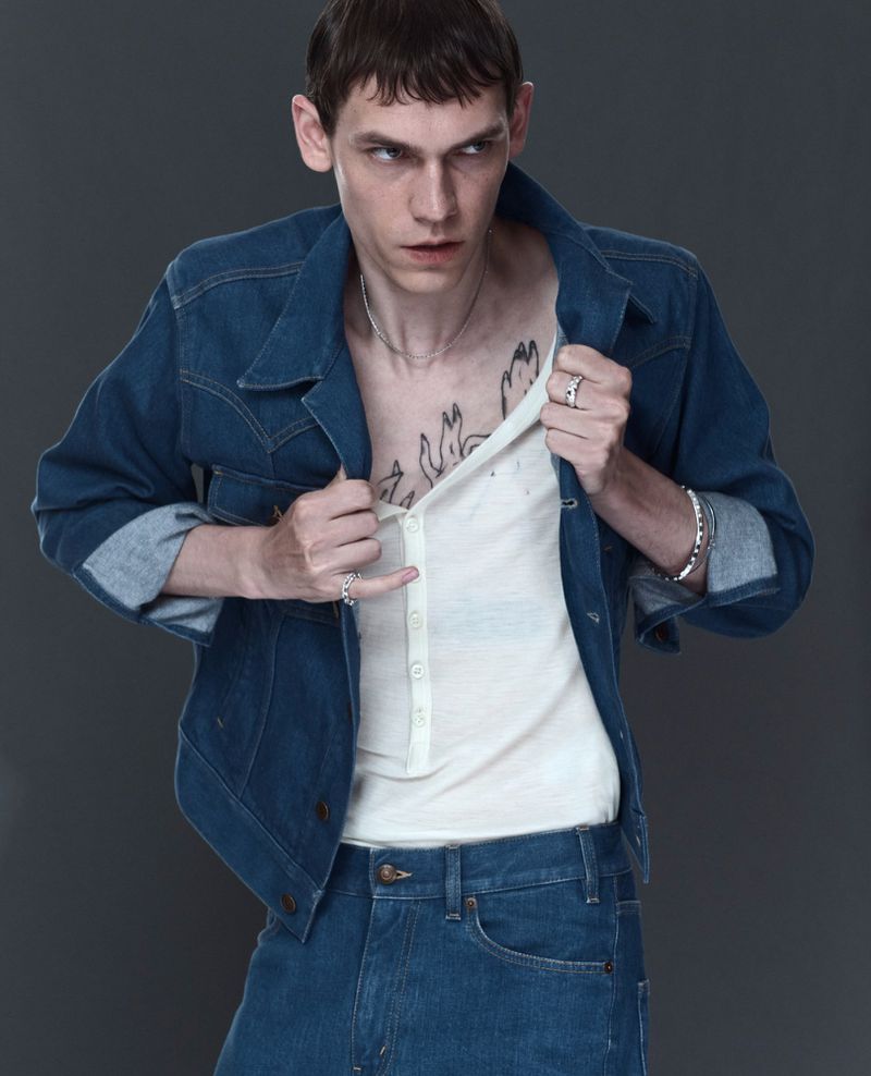 Portrait of an adult male wearing a denim jacket and jeans, posing in a studio with a plain grey background and medium shot framing.