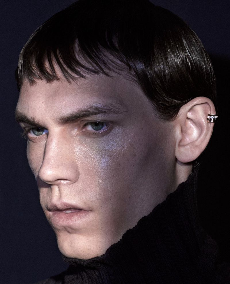Close-up portrait of a young adult man with dark hair and a black turtleneck, wearing an ear cuff, against a dark background.