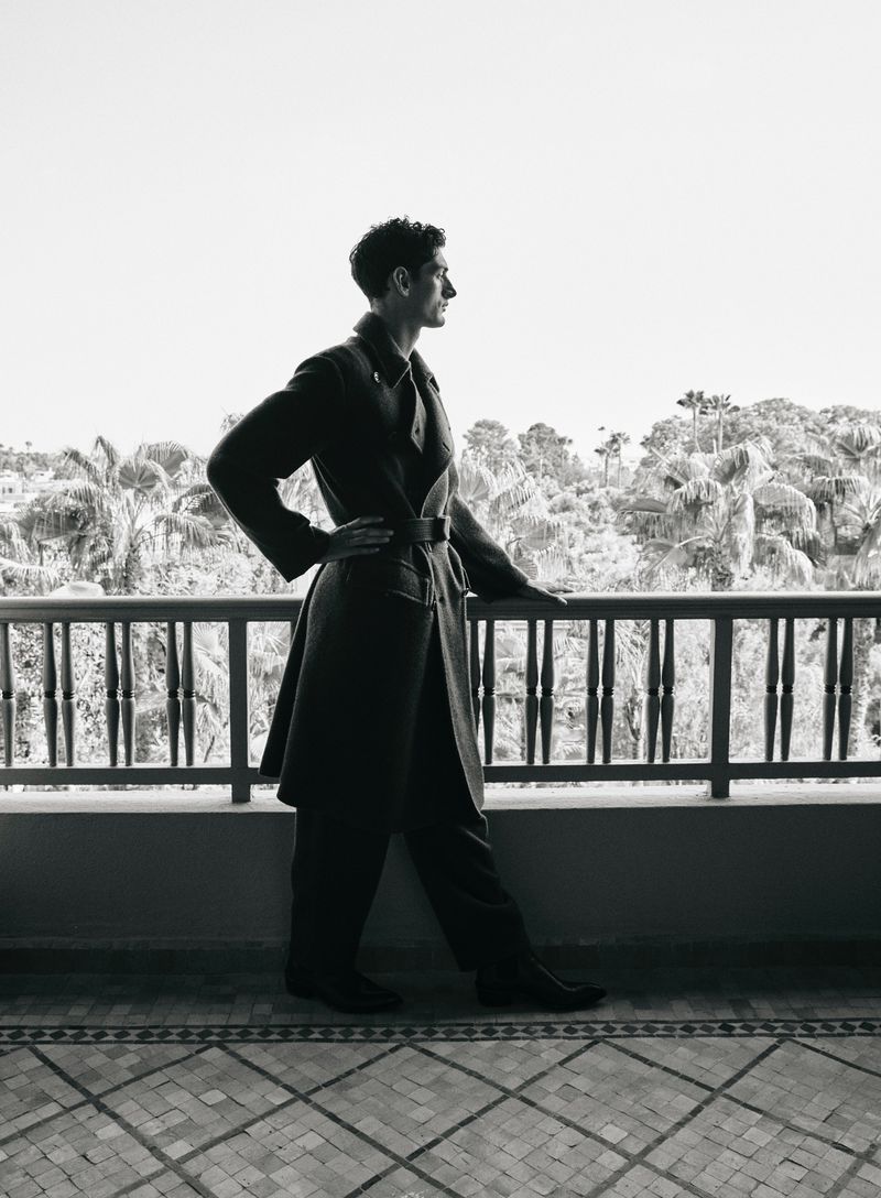 Monochrome portrait of a man in a coat and pants posing on a terrace with a background of trees and sky, captured at eye level.