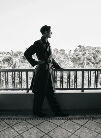 Monochrome portrait of a man in a coat and pants posing on a terrace with a background of trees and sky, captured at eye level.