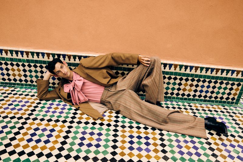 An adult male model lies on a decorative tile surface in a tan suit and pink collared shirt in a brightly lit indoor setting.