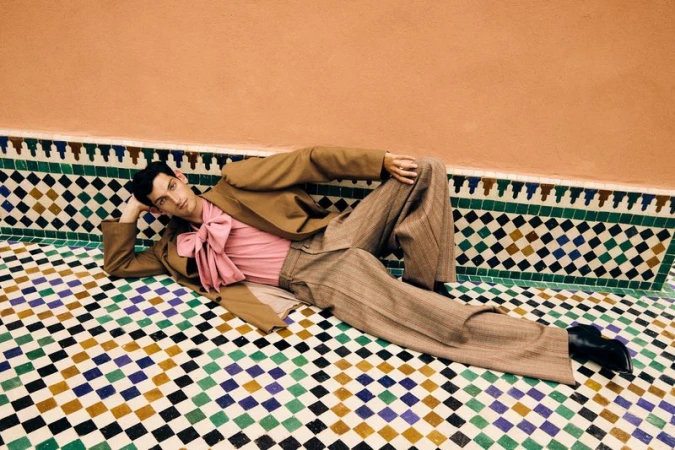 An adult male model lies on a decorative tile surface in a tan suit and pink collared shirt in a brightly lit indoor setting.