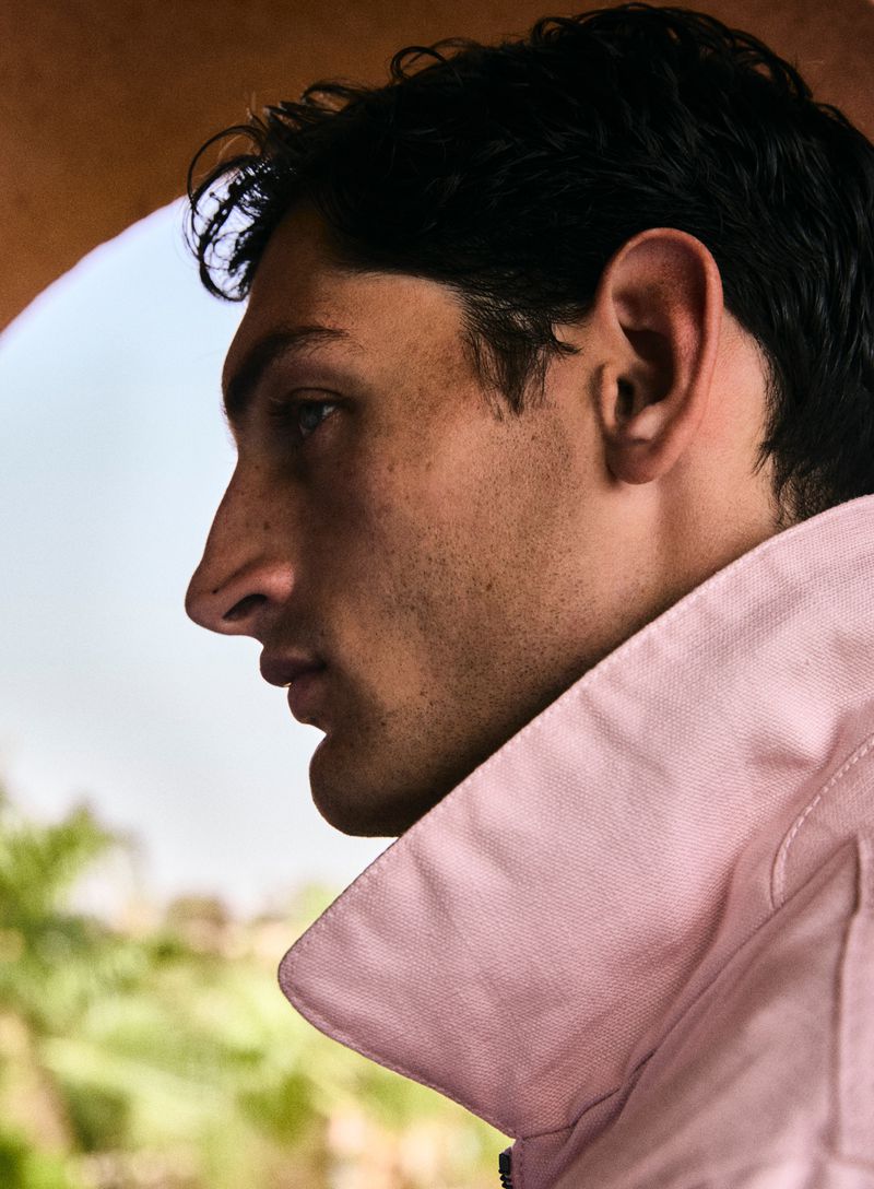 Profile of a young adult man with dark hair, wearing a pink collared shirt, posing outdoors under a stone archway with greenery visible.