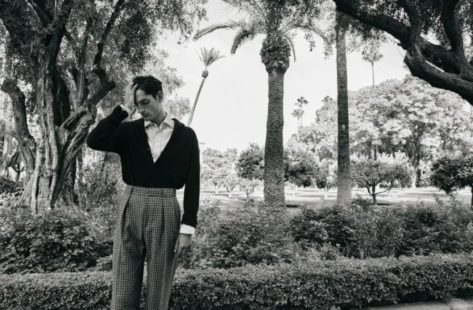 Model Aaron Shandel posing with hand to head wearing a dark cardigan and high-waisted trousers in an outdoor park.