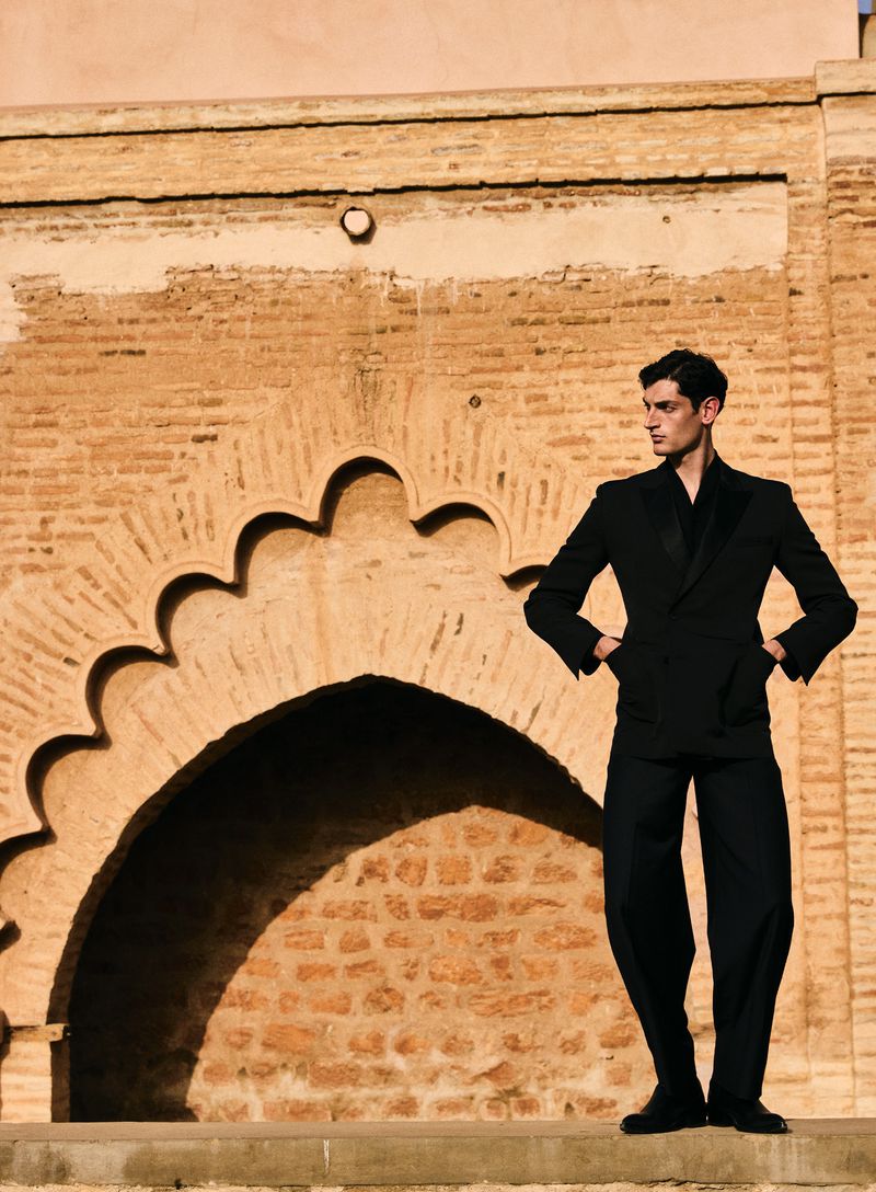 A stylish man in a black suit stands against an adobe wall with an archway in warm sunlight, captured in a fashion-forward editorial shot.