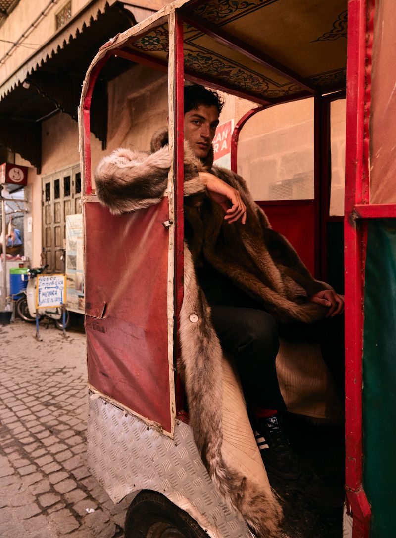 An adult male sits inside a red, green, and white auto rickshaw on a cobblestone street. He wears a fur coat and black pants.