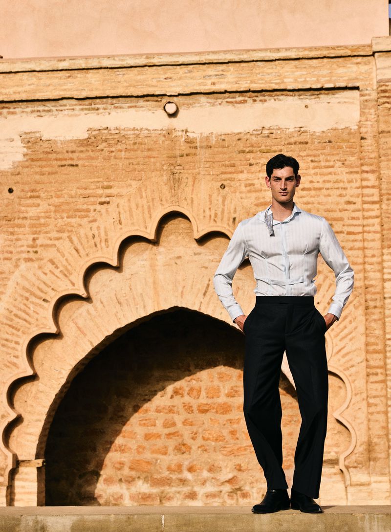 A full shot of a young adult male model posing in a light blue dress shirt and black dress pants in front of a patterned brick wall.