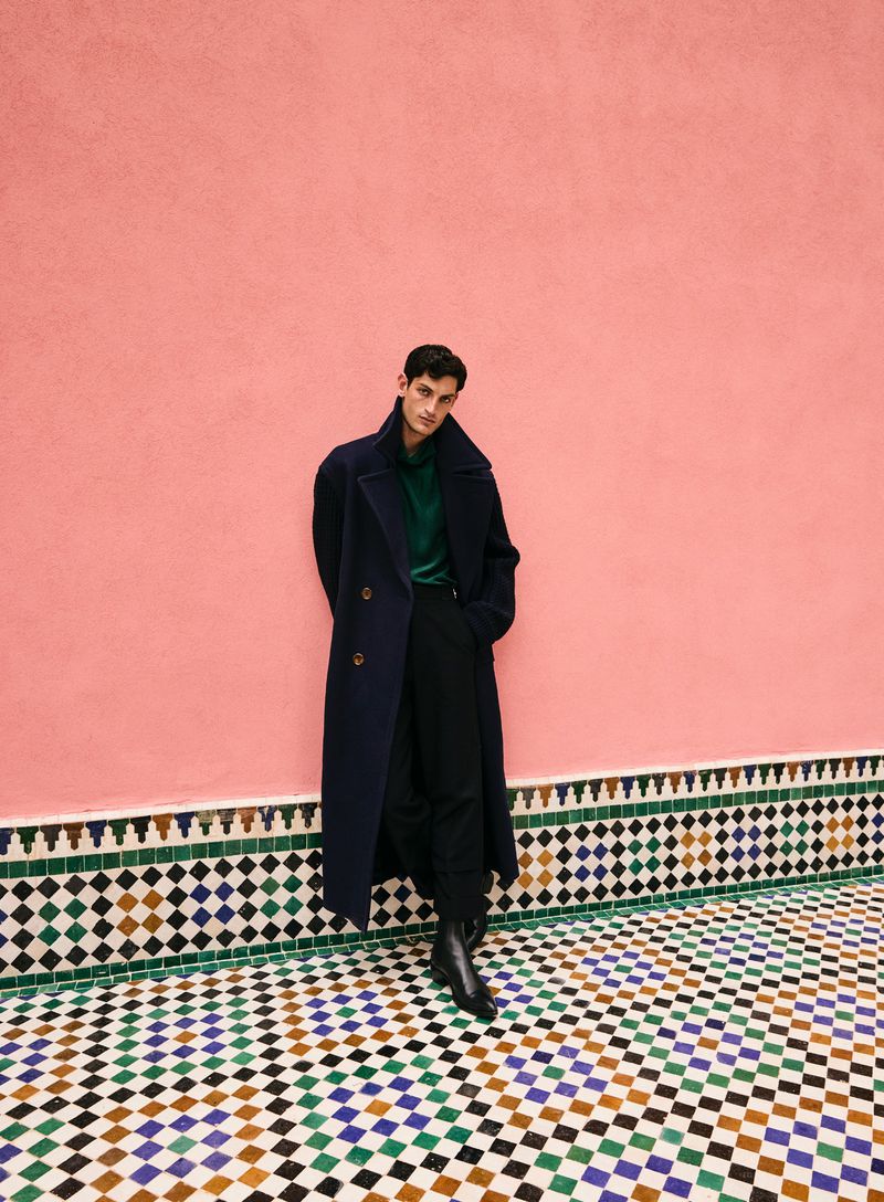 A male model poses in a dark overcoat and black trousers against a pink wall with decorative tiling on the lower wall and floor.