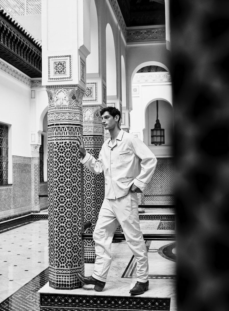 Monochrome portrait of a man in pajamas posing against an ornately tiled column in a tiled courtyard with arches and decorative details.