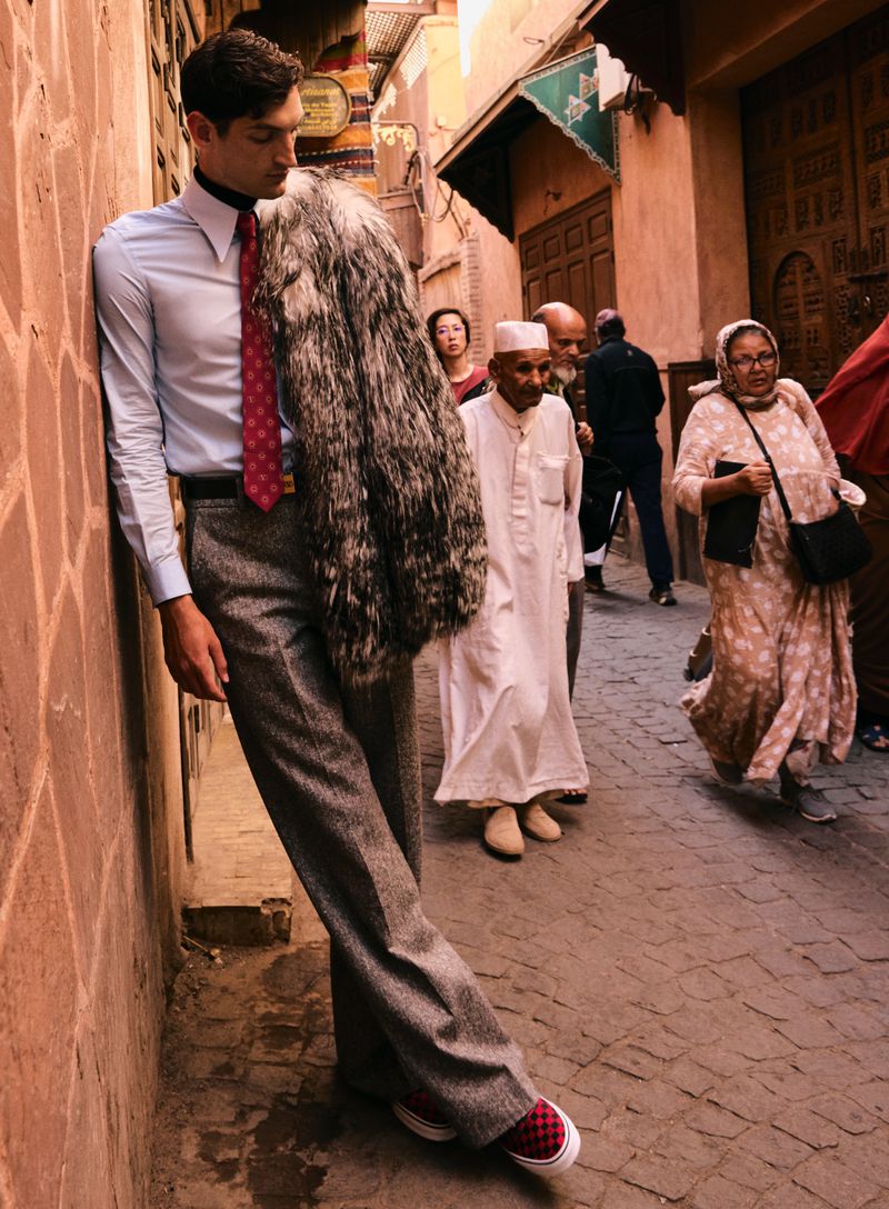Male model in an azure dress shirt, tie, and grey tweed pants leans against a textured orange wall in a city street setting.