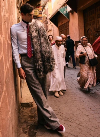 Male model in an azure dress shirt, tie, and grey tweed pants leans against a textured orange wall in a city street setting.