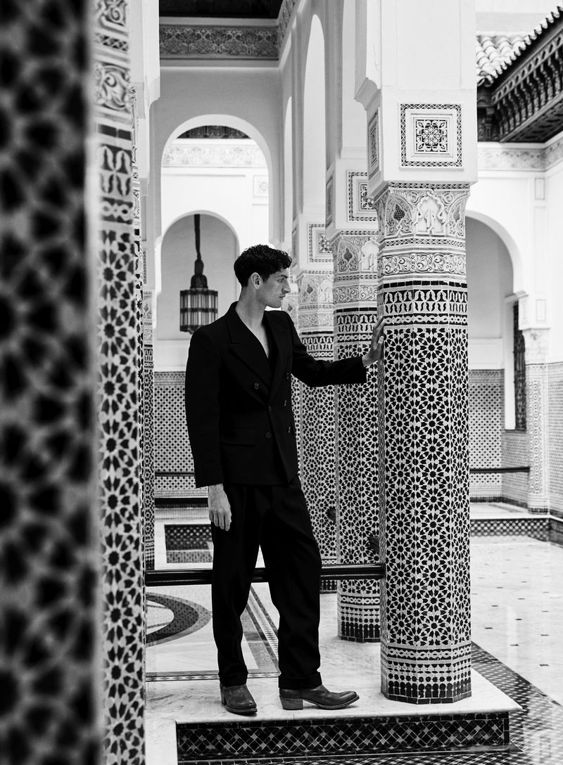 Man in a black suit poses indoors among pillars with intricate tile patterns, creating a stylish and timeless fashion image.