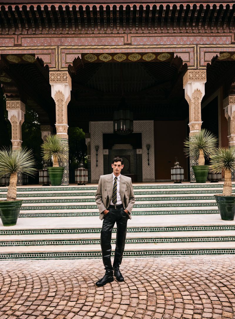 Man in suit and tie standing in front of building with arches and potted plants, wearing leather pants and boots on stone pavers.