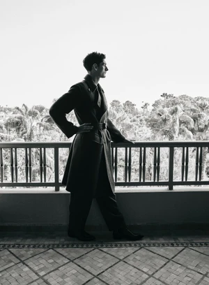 Model Aaron Shandel posing in profile on a terrace wearing a belted overcoat in black and white for GQ España.