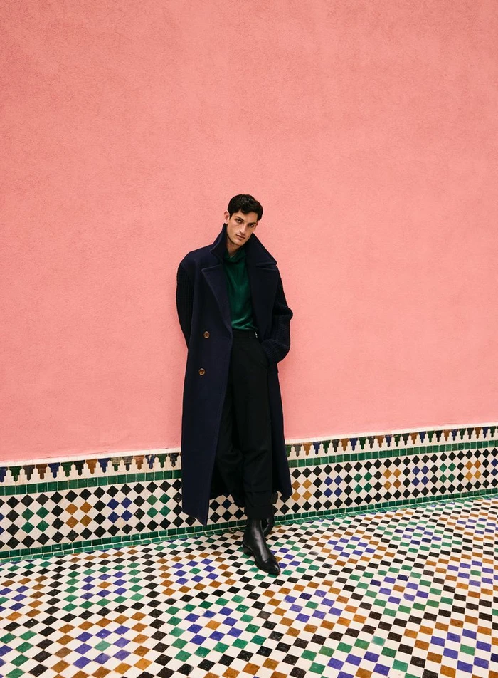 Man wearing a long navy coat leaning against a pink wall above a mosaic-tiled floor.