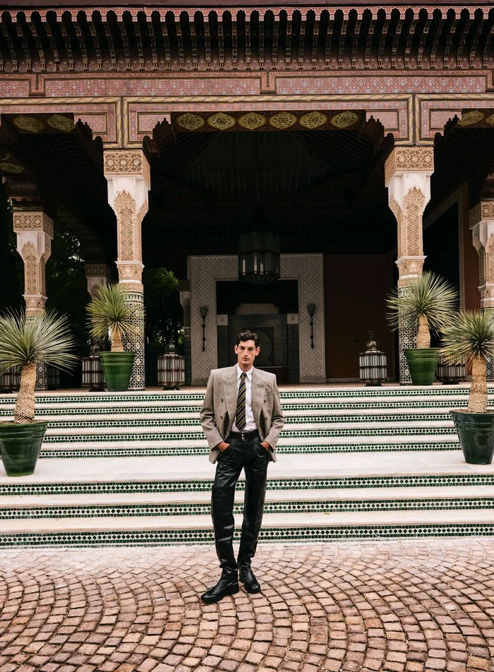 Model Aaron Shandel standing in a beige double-breasted blazer and black leather pants in front of an ornate tiled pavilion.