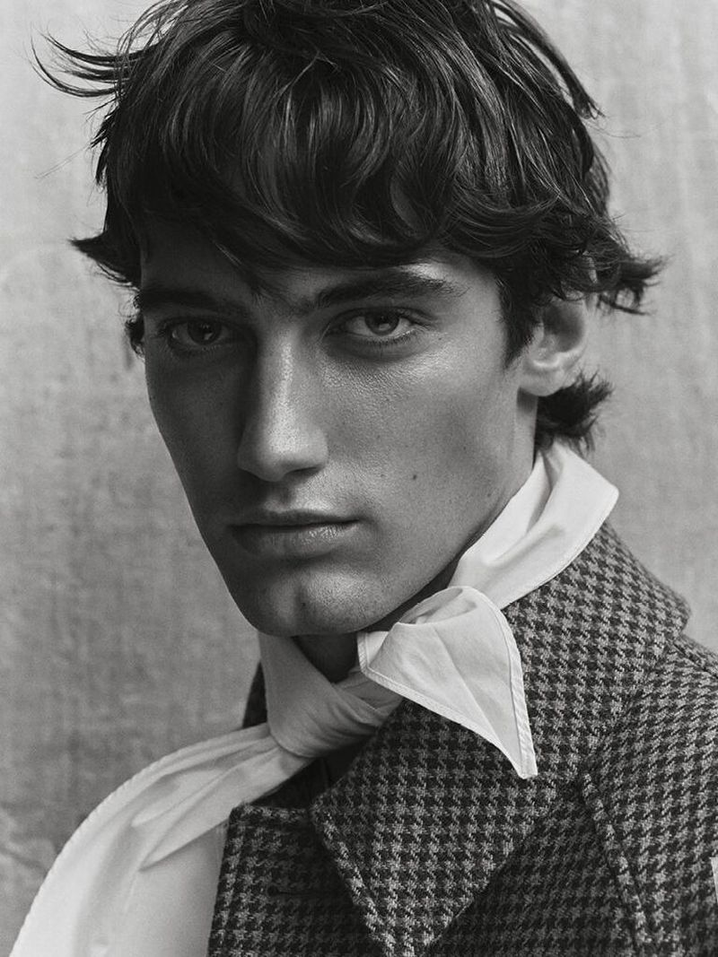 Portrait of a young adult male with dark hair wearing a patterned coat and a white scarf, set against a neutral background.