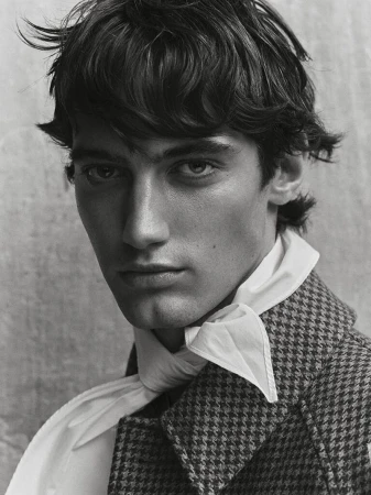 Portrait of a young adult male with dark hair wearing a patterned coat and a white scarf, set against a neutral background.