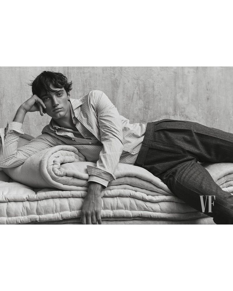 Monochrome portrait of a young adult man reclining on a quilted blanket in a striped shirt and dark pants in front of a concrete wall.