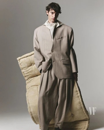 A male model poses in an oversized beige suit next to a large floor cushion in a studio setting for a fashion editorial shot.