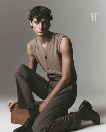 Model kneeling on a wooden crate wearing a beige ribbed sleeveless knit vest and brown trousers against a grey backdrop.