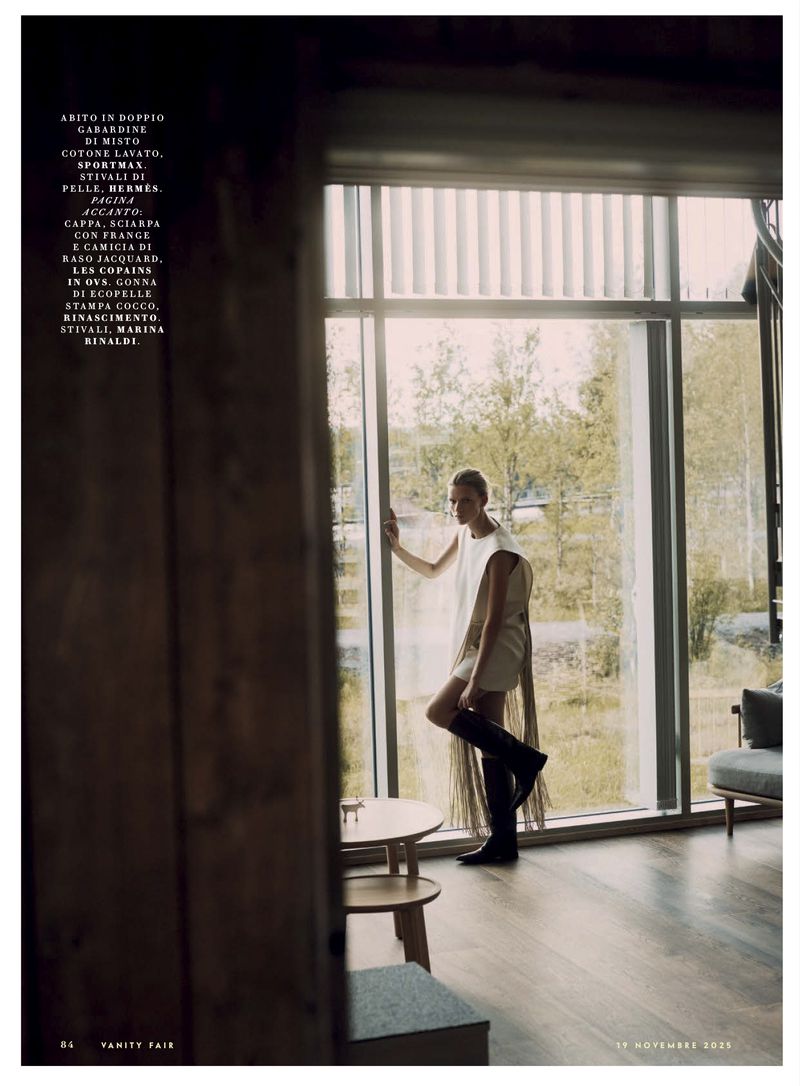 Model standing by a large window in a light dress and black boots, viewed through a doorway for Vanity Fair Italia