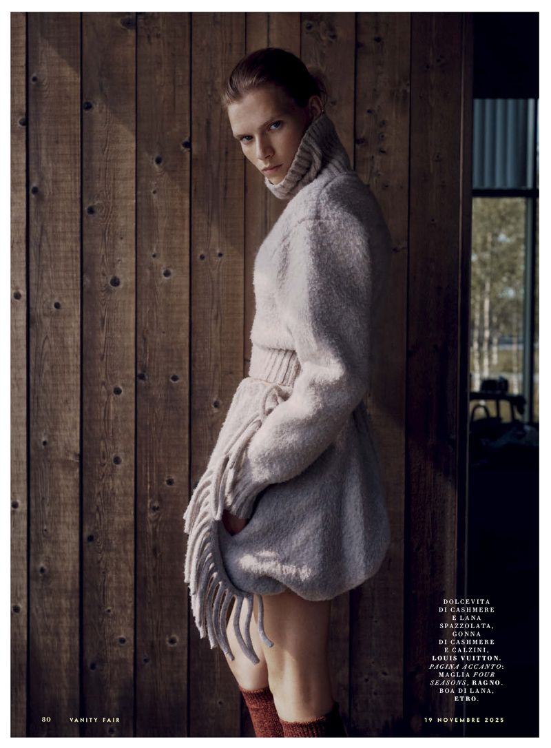 A woman poses indoors by a wooden wall, wearing a soft grey wool sweater, knit skirt with fringe, and wool socks, in diffused light.