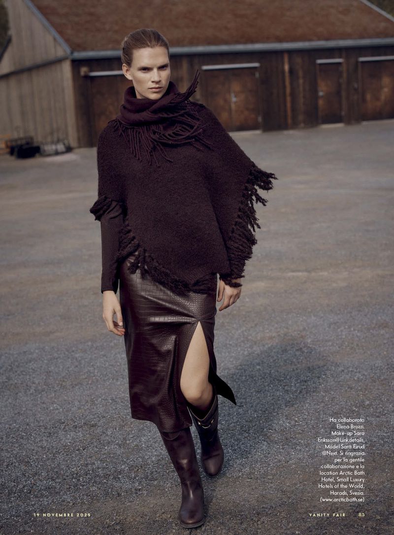 A woman in a brown fringed poncho, brown snakeskin skirt, and boots poses in front of a barn for a fashion editorial.