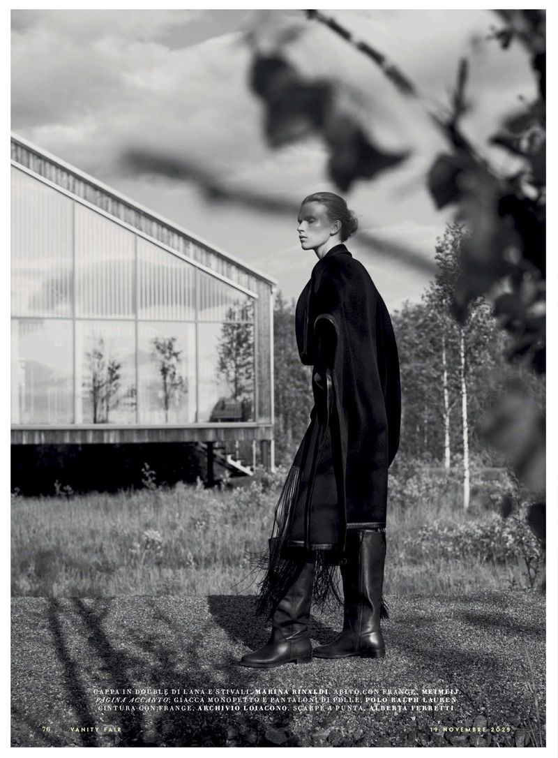 A woman in dark clothing poses outdoors near a modern house with glass walls, against a backdrop of trees in a black and white fashion shot.