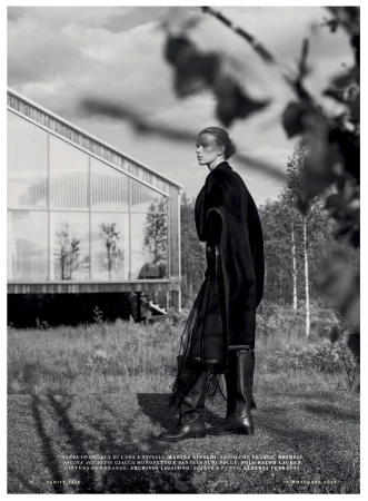 A woman in dark clothing poses outdoors near a modern house with glass walls, against a backdrop of trees in a black and white fashion shot.