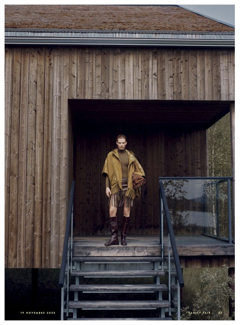 An adult woman standing on wooden steps, wearing a poncho with fringe, a brown top, and dark boots, posed in front of a wooden building.