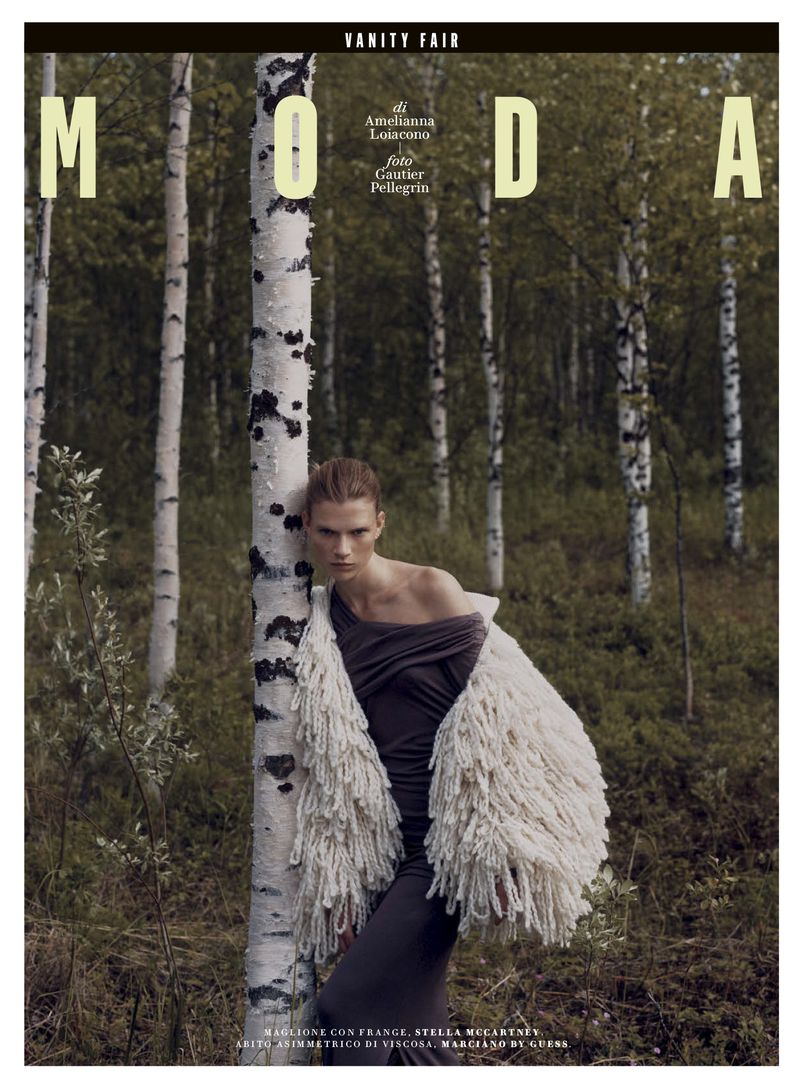 Fashion magazine cover: a woman in a draped dress and shaggy coat leans against a birch tree in a birch tree forest setting.