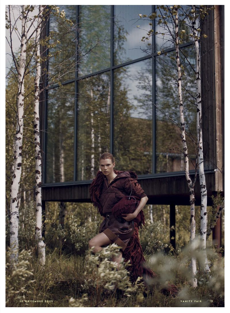 Fashion editorial shot of a woman in a textured outfit posing outdoors near birch trees and a modern home with large reflective windows.