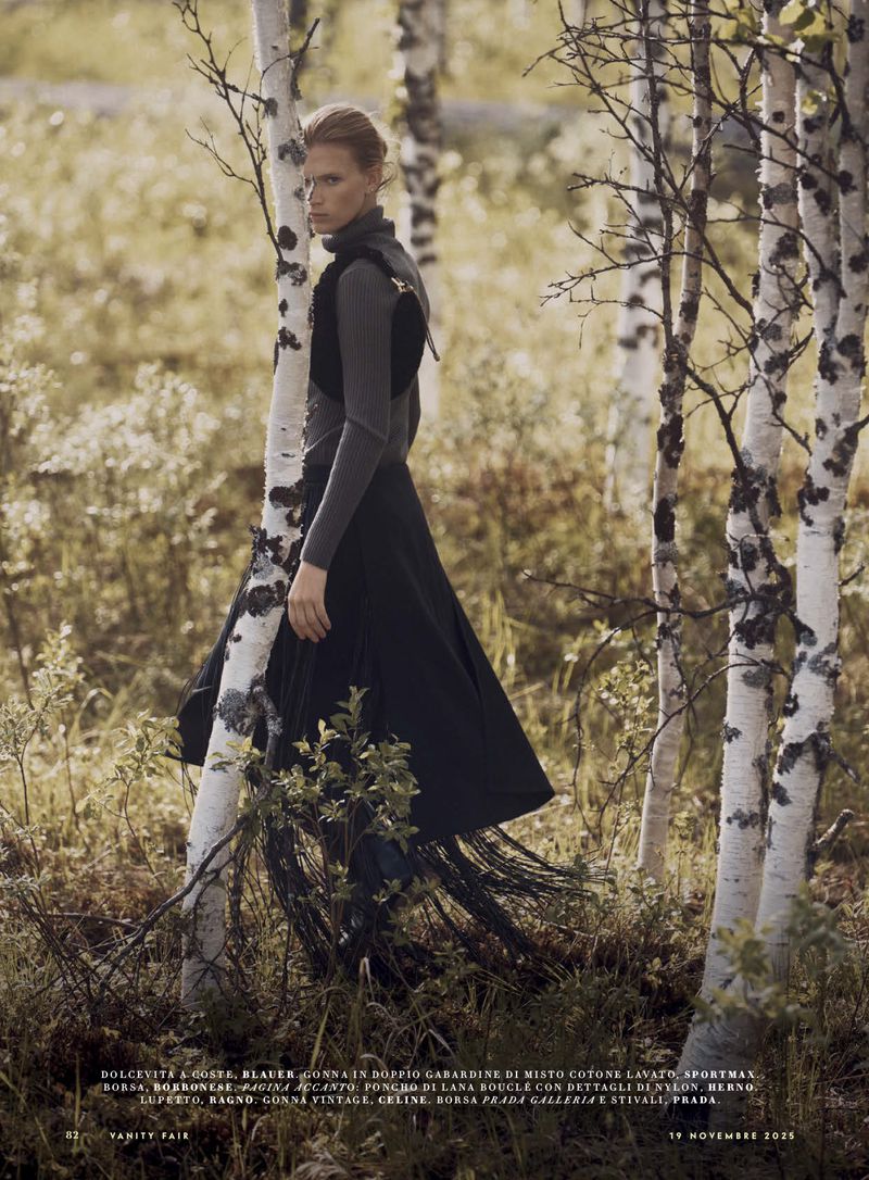 Fashion editorial shot of a woman in a gray turtleneck and long black fringe skirt standing in a birch tree forest setting.